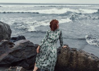 Red-haired woman in a patterned dress sits by the rocks on a beach looking at the ocean waves.
