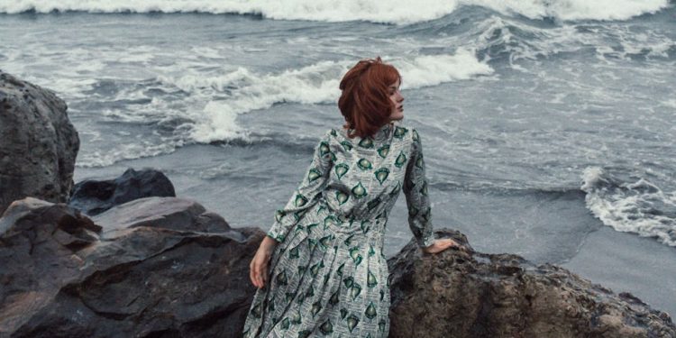 Red-haired woman in a patterned dress sits by the rocks on a beach looking at the ocean waves.