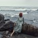 Red-haired woman in a patterned dress sits by the rocks on a beach looking at the ocean waves.