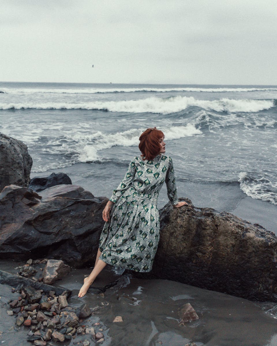 Red-haired woman in a patterned dress sits by the rocks on a beach looking at the ocean waves.