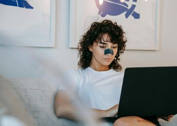 Focused young female freelancer with curly hair in casual outfit sitting on sofa with anti acne patch on nose and working remotely on laptop