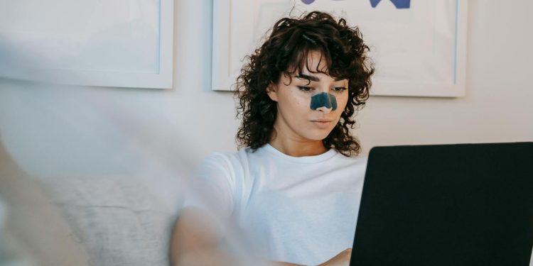 Focused young female freelancer with curly hair in casual outfit sitting on sofa with anti acne patch on nose and working remotely on laptop