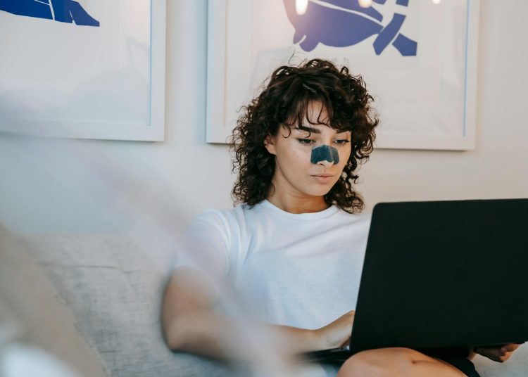 Focused young female freelancer with curly hair in casual outfit sitting on sofa with anti acne patch on nose and working remotely on laptop