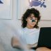Focused young female freelancer with curly hair in casual outfit sitting on sofa with anti acne patch on nose and working remotely on laptop